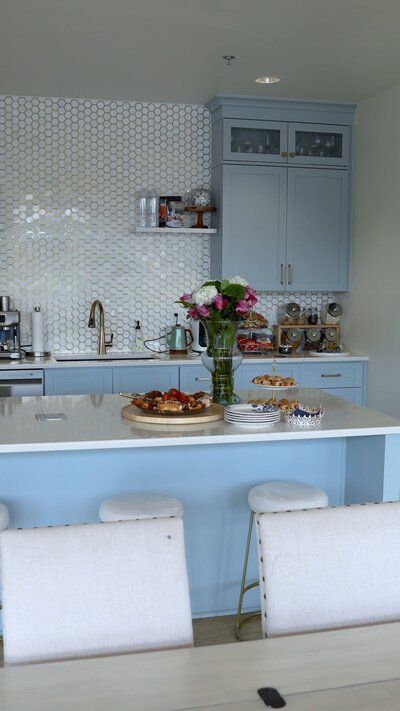 Light blue kitchen with a white countertop and island, featuring cabinets, a flower bouquet, and bar stools.
