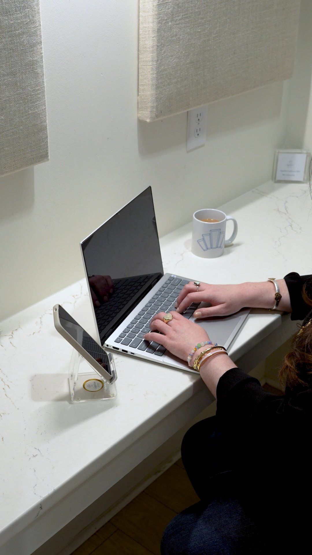 Person typing on a laptop at a white desk; a mug and phone are present.