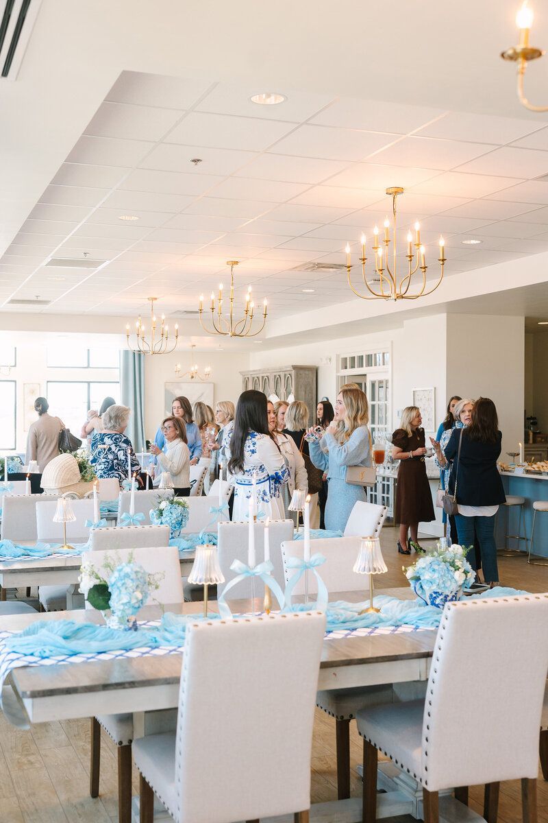 A light-filled room with tables set for an event. People in light-colored clothes mingle. Centerpieces include blue flowers.