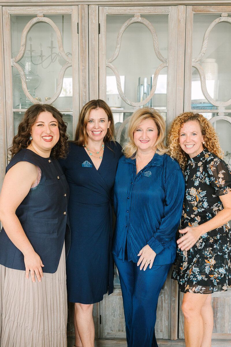 Four women stand smiling together in front of a light-colored, ornate glass-paneled display cabinet.