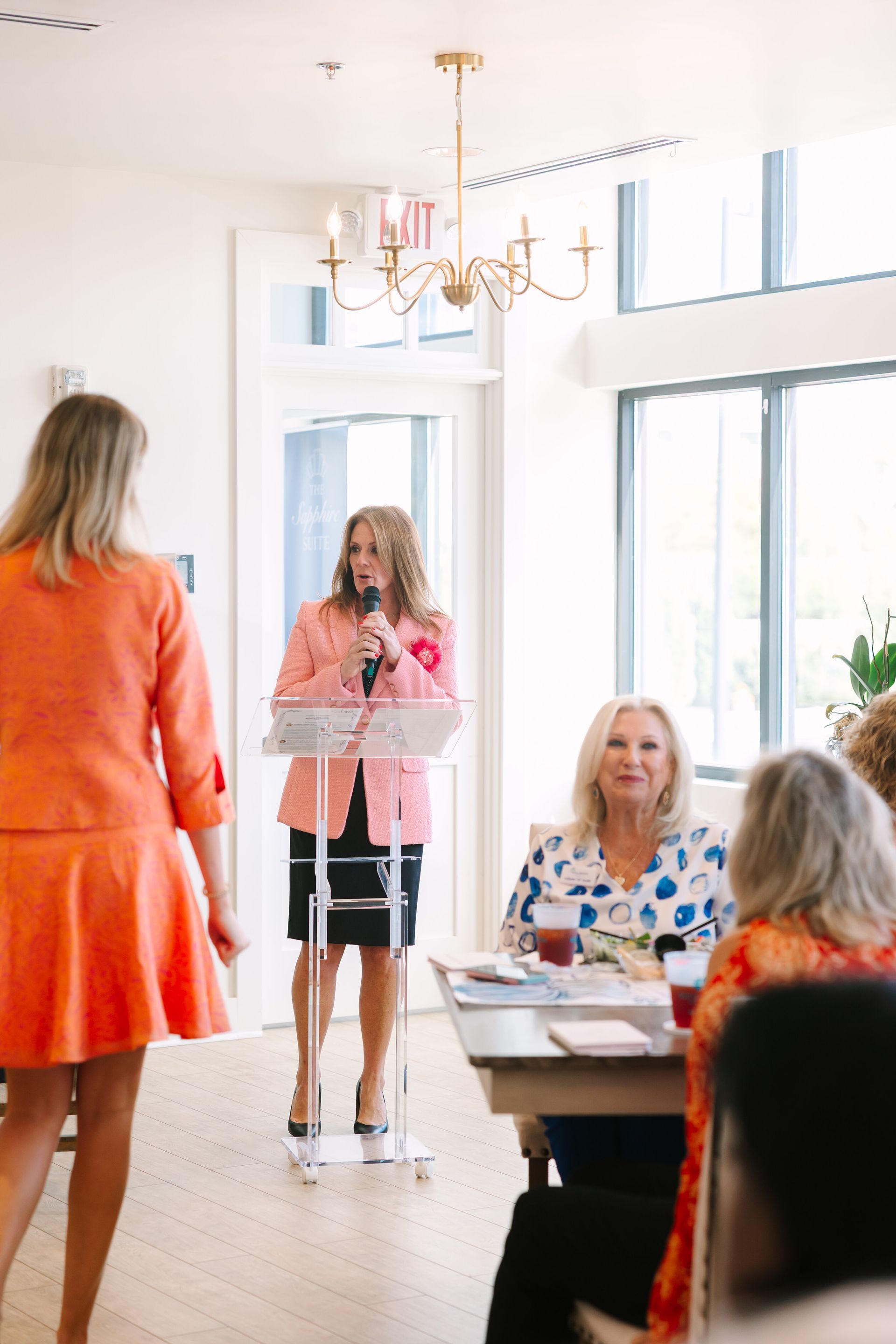 Woman speaking at a podium in a light-filled room. Another woman walks past, and others are seated at a table.