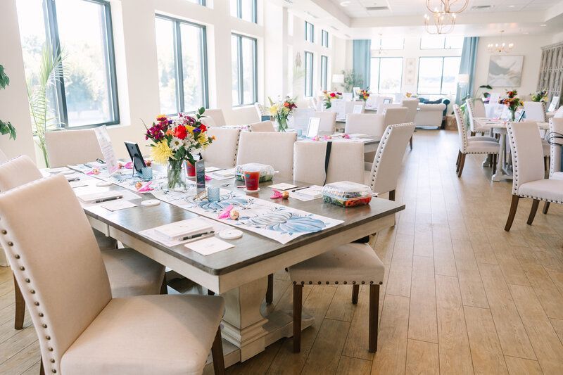 Dining room with tables set for an event; light-colored chairs, floral centerpieces, windows.