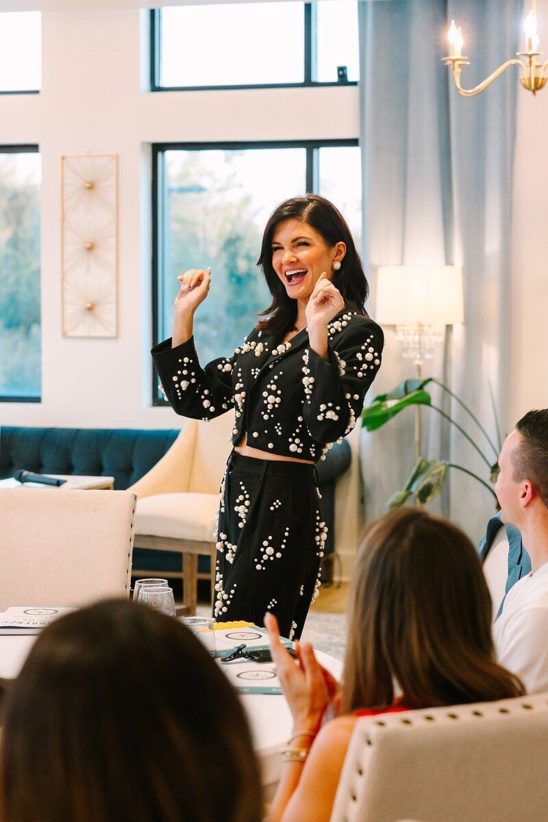 Woman in black and white outfit gestures excitedly. People at a table. Indoor setting with large window.