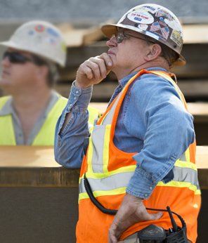 Construction worker in hard hat and safety vest, contemplating. Another worker in background.