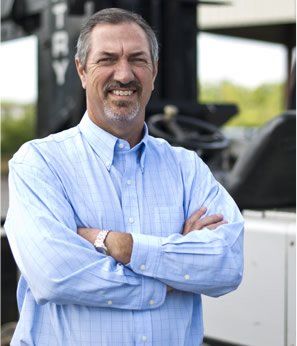 Man with crossed arms, smiling, wearing a blue shirt, standing outdoors in front of machinery.