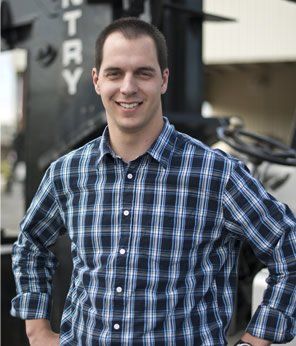 Man in plaid shirt smiles, hands on hips, in front of a large machine outdoors.