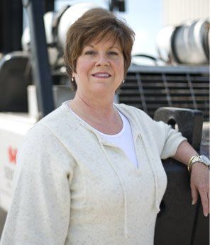 Woman in a cream sweater smiles, leaning on machinery, with a white shirt underneath; outdoor setting.