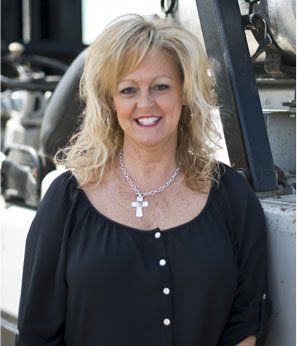Woman with blonde hair smiles in front of machinery, wearing a black shirt and cross necklace.