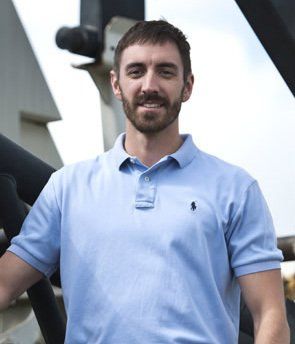 Man in light blue polo shirt smiles, standing near metal machinery.