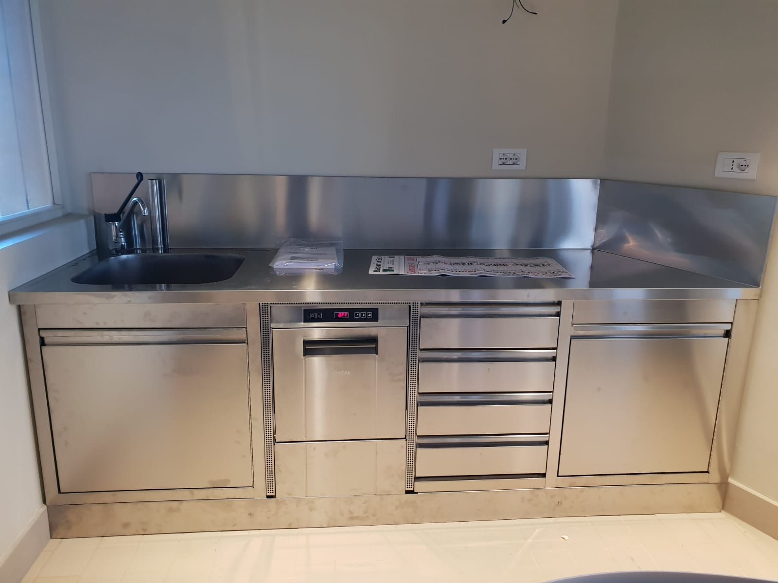 Piccola cucina con piano di lavoro, lavello, forno e cassetti in acciaio inox, addossata a una parete chiara.