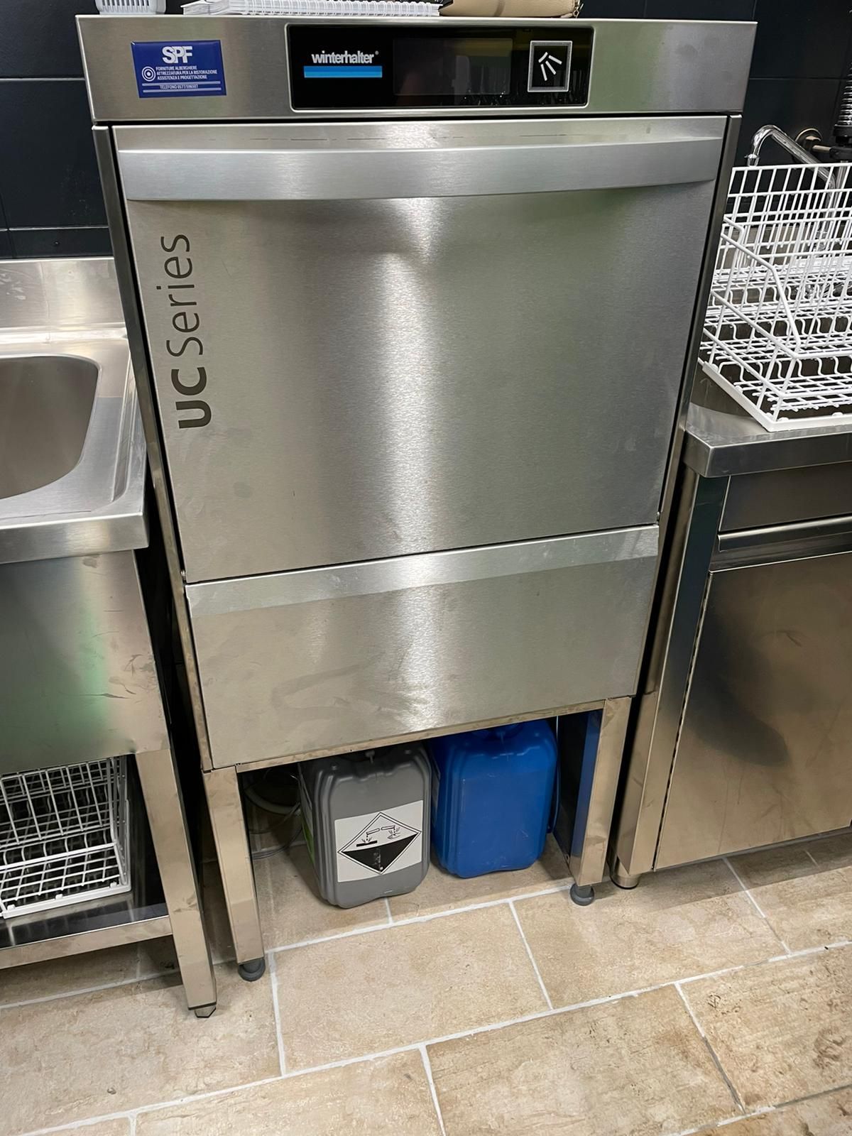 Lavastoviglie professionale serie UC in acciaio inox in una cucina, con i bidoni della spazzatura riposti al di sotto.