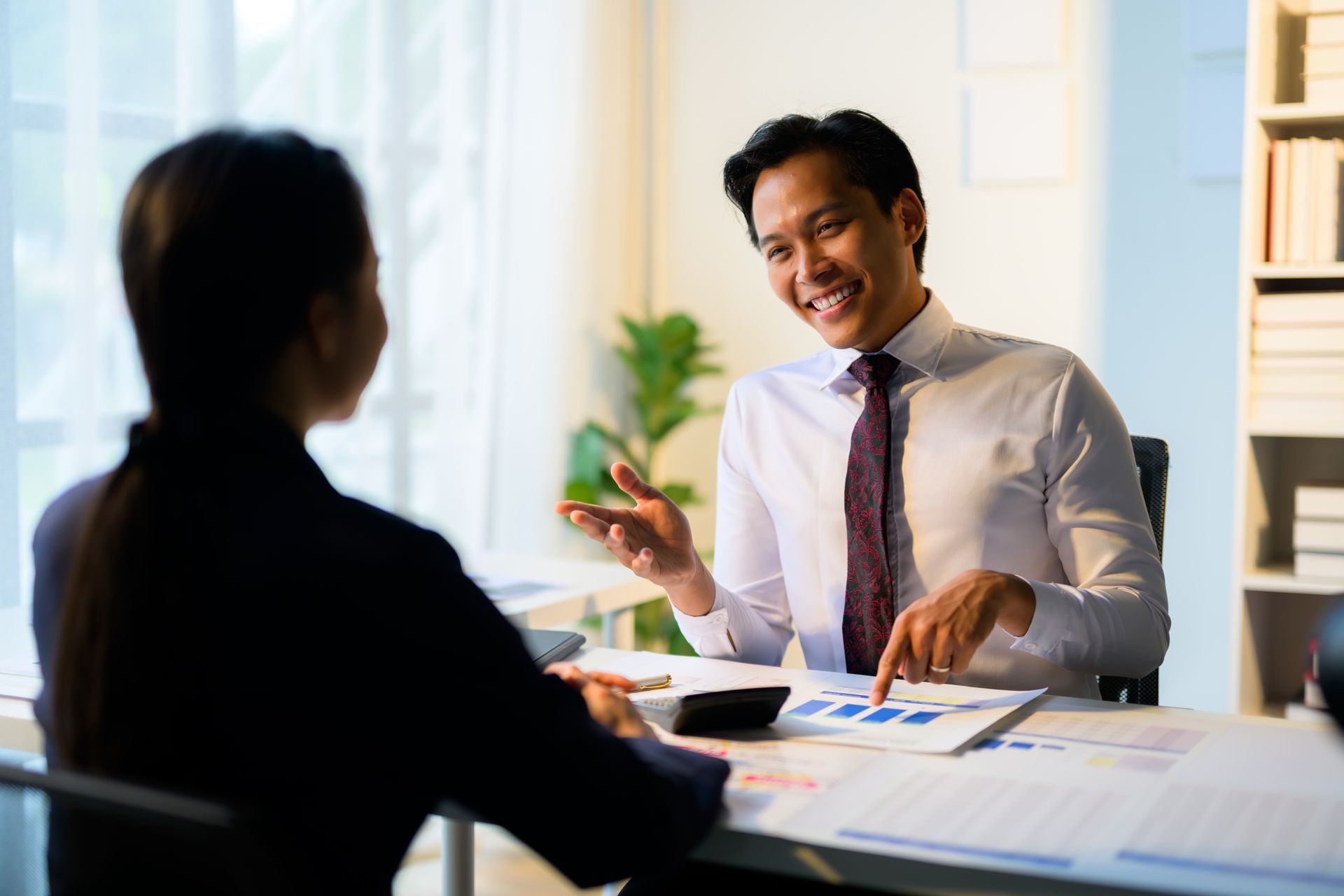 Man in white shirt gestures at a document while speaking to a person at a desk.