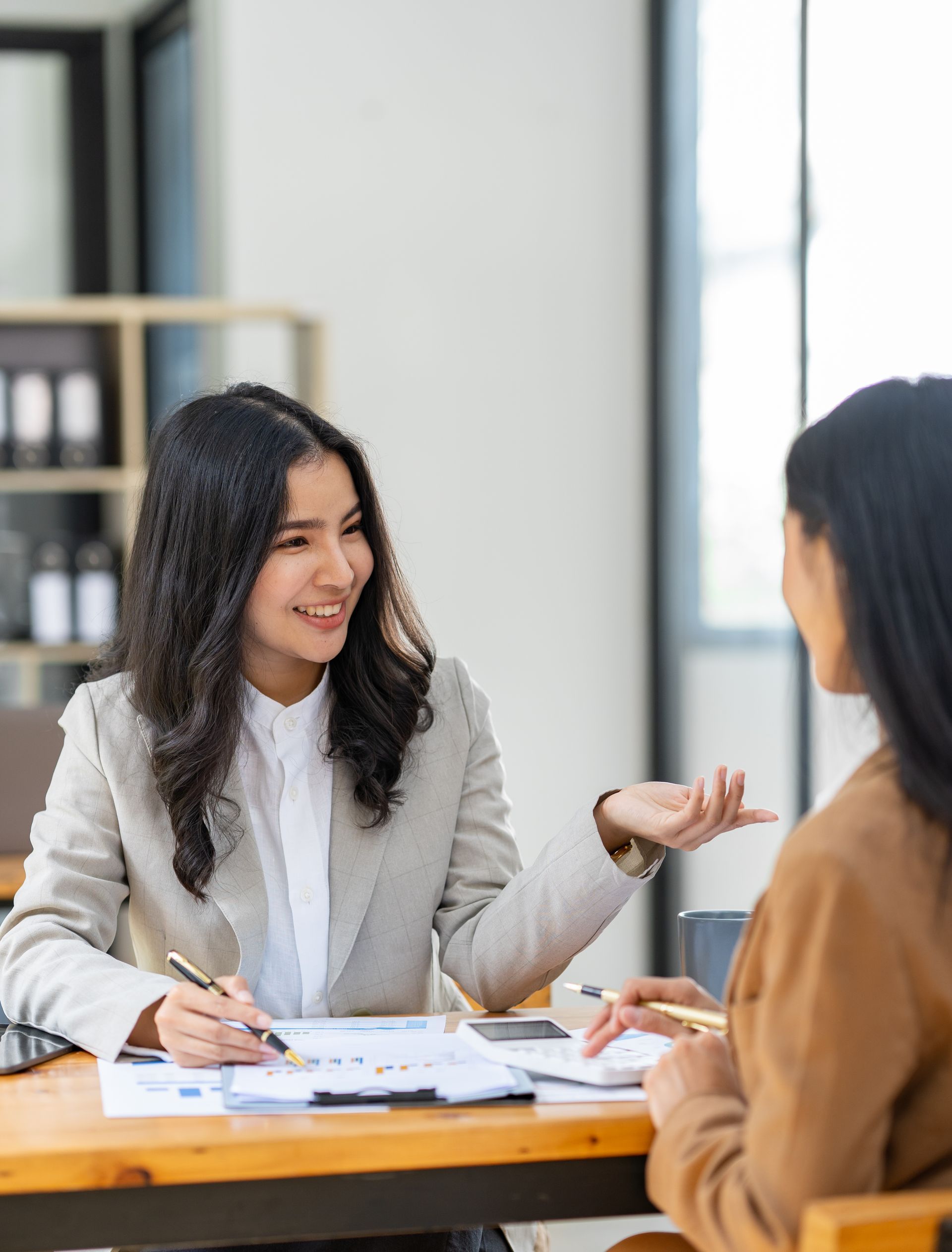 Woman in blazer gestures, speaking to another woman at a desk with papers, in an office.