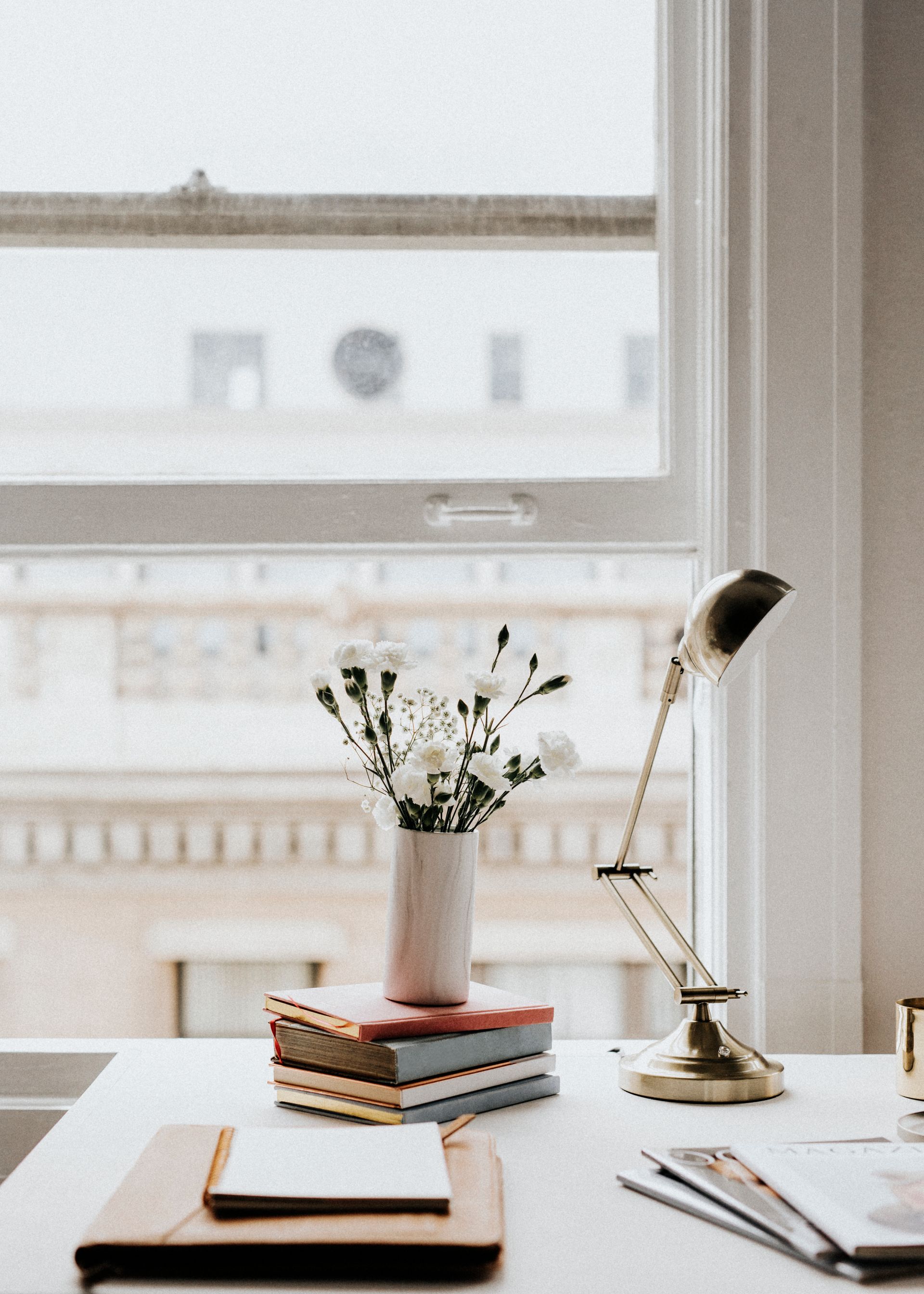 Desk with flowers, books, lamp, and notepad by a window, with an exterior view.