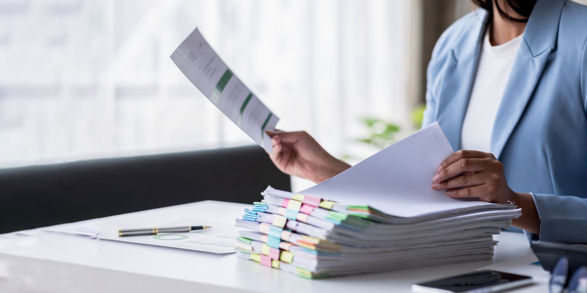 Person in a blue blazer reviewing a stack of papers at a desk with a pen and phone.