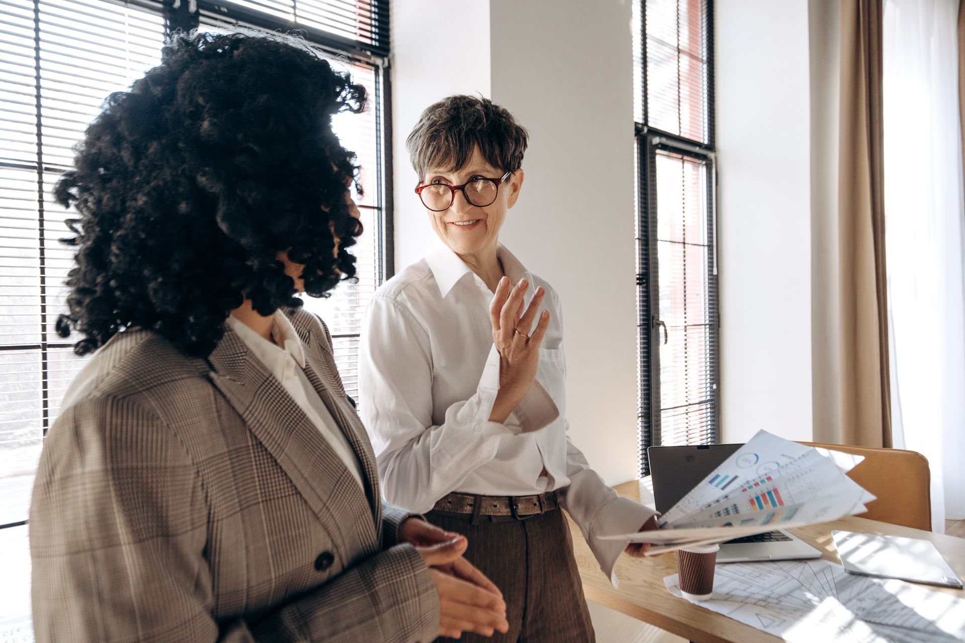 Two women at a desk reviewing documents with laptops, calculator, and pen, focused discussion.