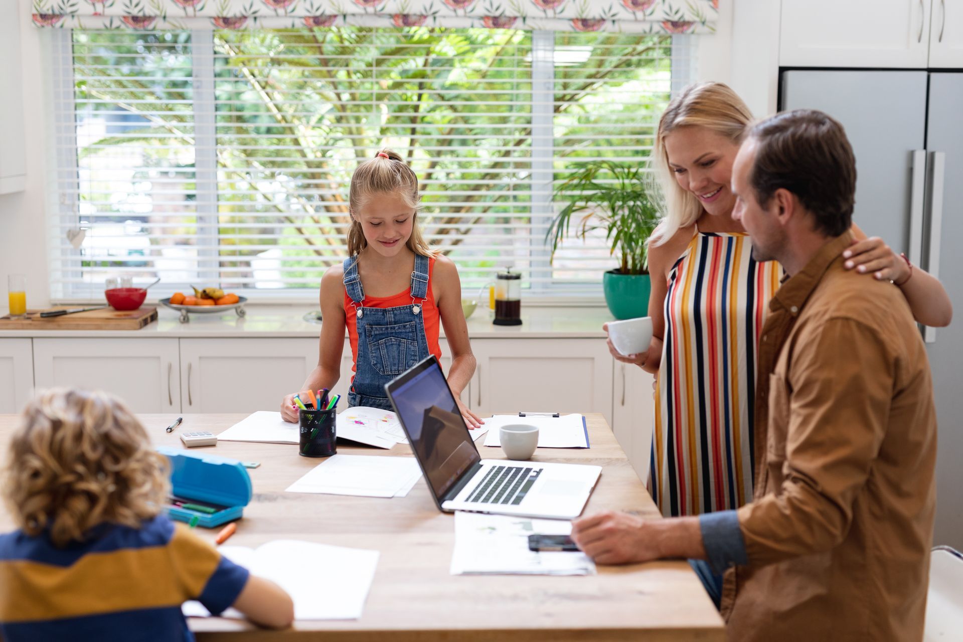 Family in a kitchen: mother with cup, father at laptop, daughter writing, son drawing. Bright natural light.