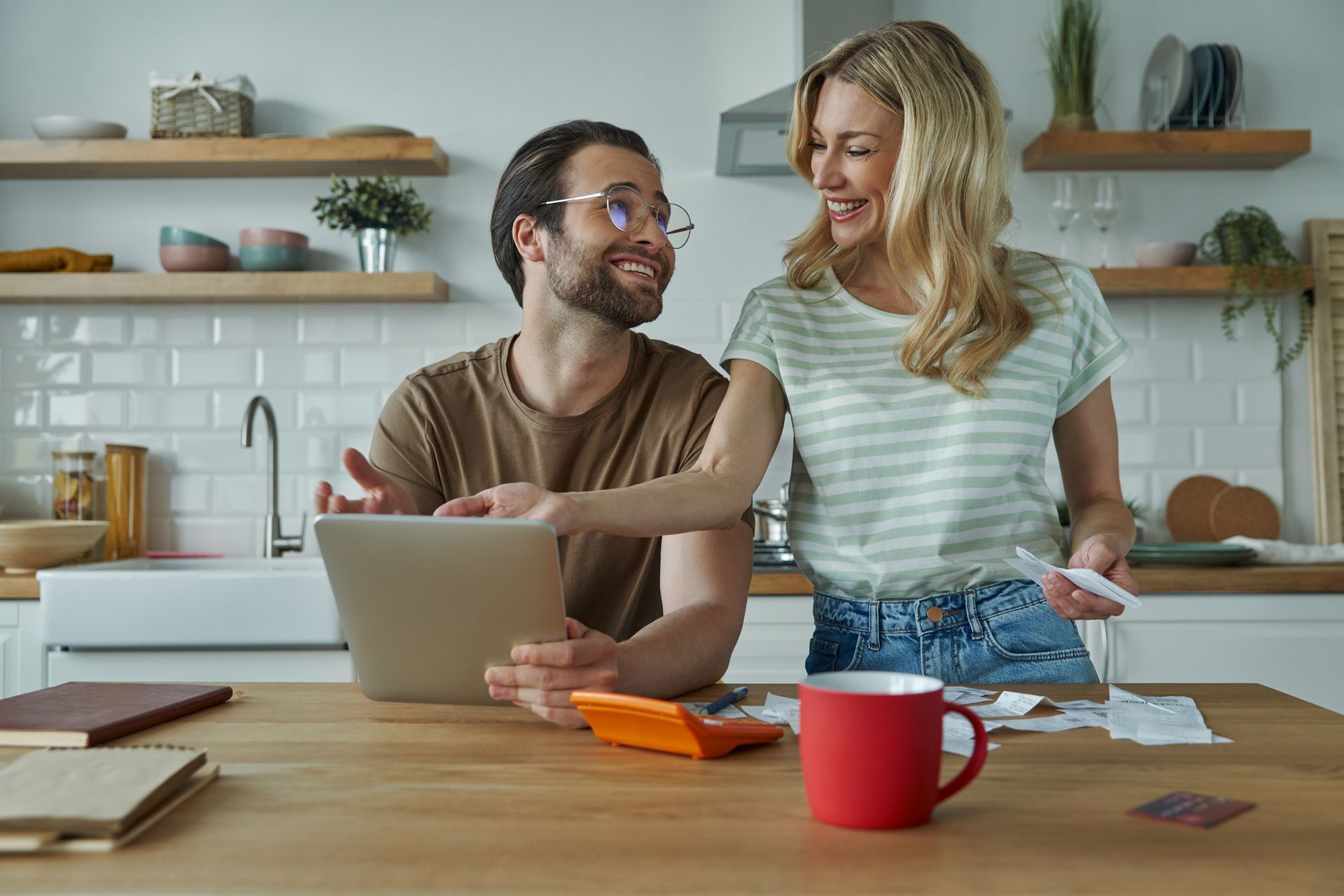 Couple in kitchen, looking at tablet, bills, and calculator, smiling.