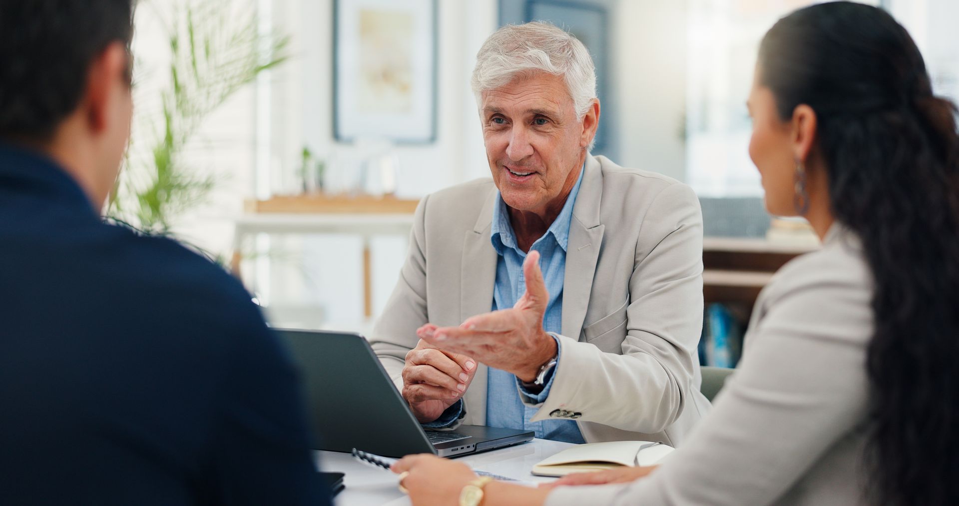 Man in blazer gestures during a meeting with two colleagues at a table with a laptop.
