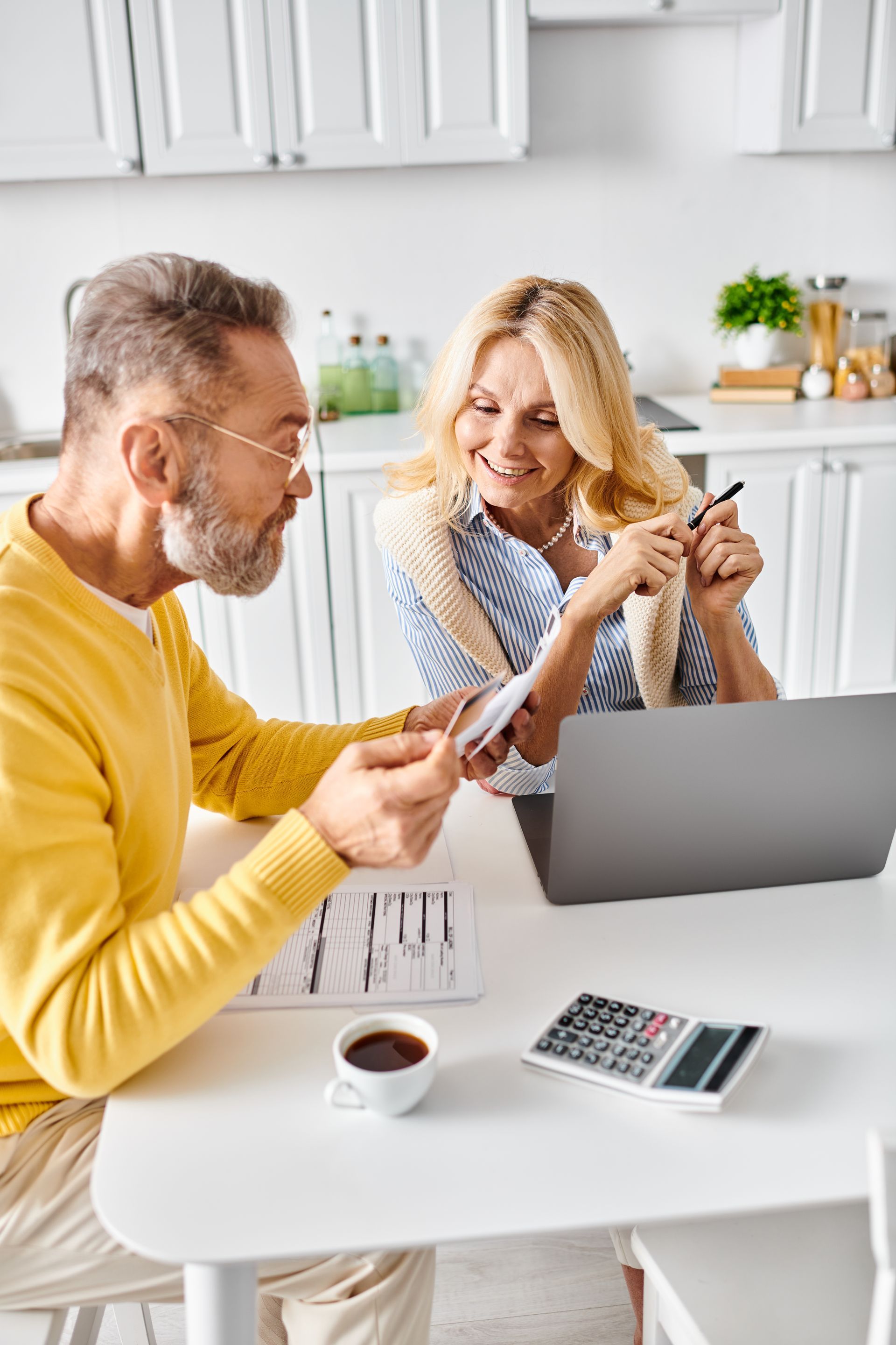 Man and woman review documents at a table with laptop, calculator, and coffee. Bright kitchen setting.