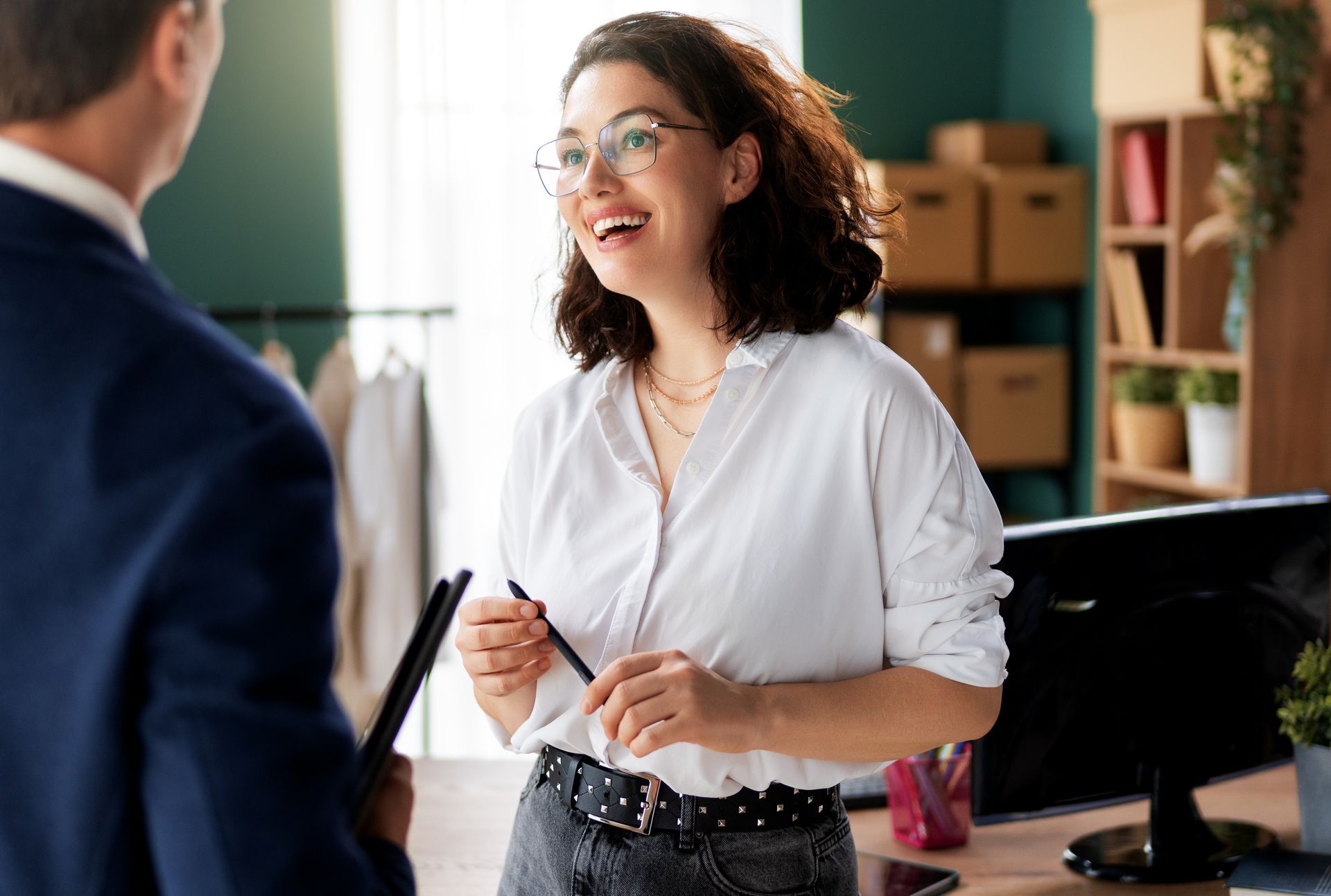 Woman in white shirt smiles, talking with person in suit in office.
