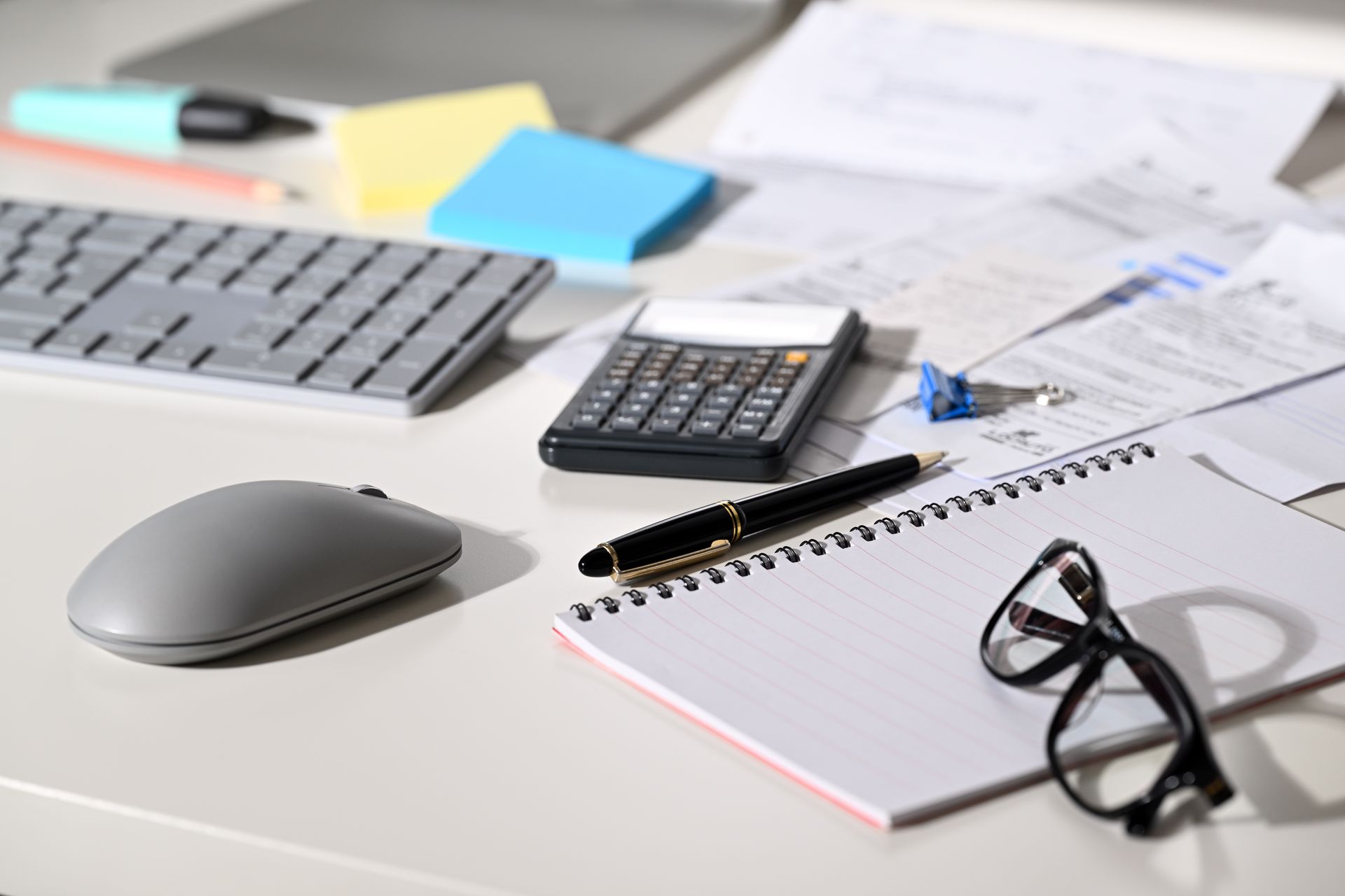 Office desk with a calculator, keyboard, notepad, glasses, and pen.