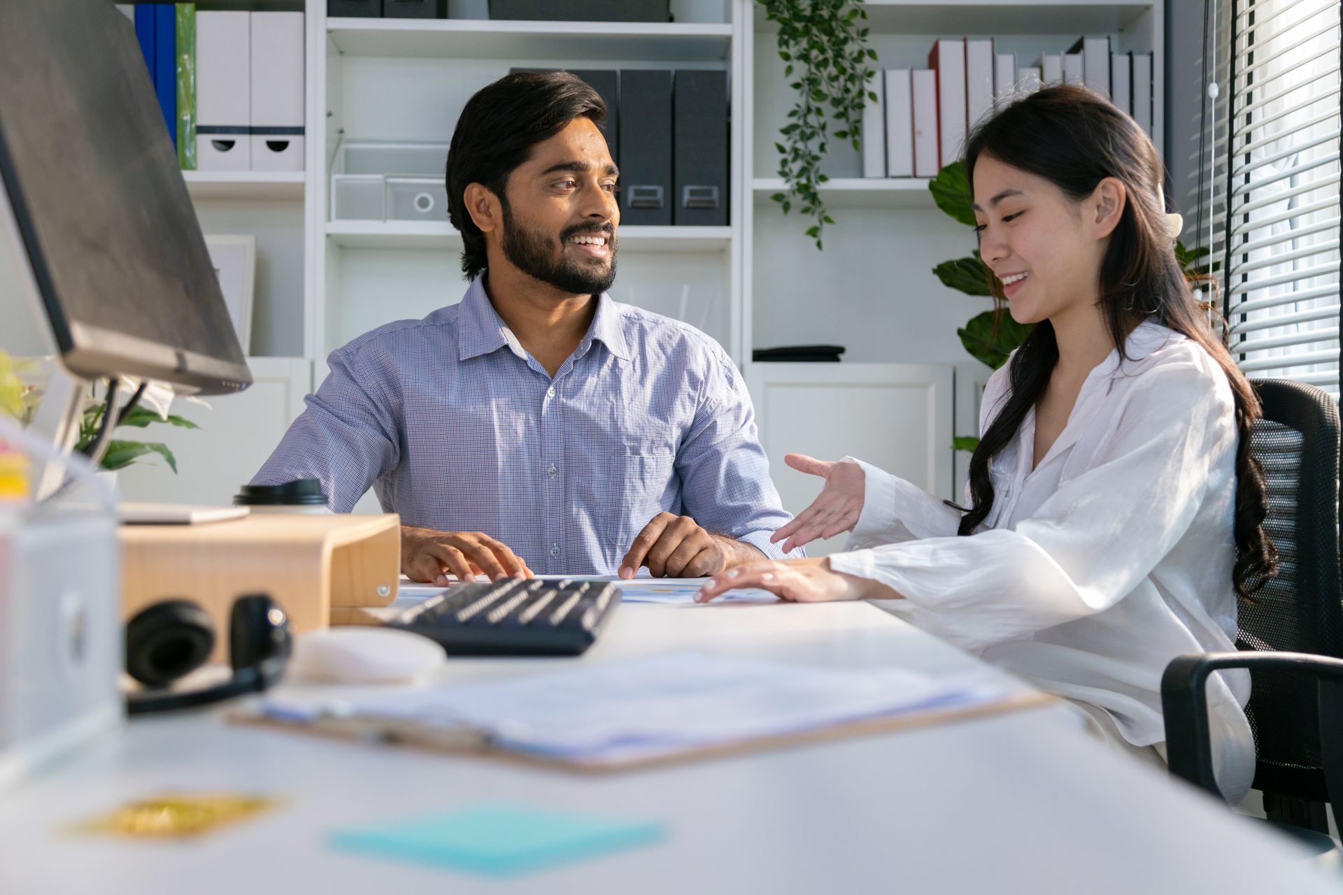 Man and woman smiling and gesturing at a desk with documents and a computer in an office setting.