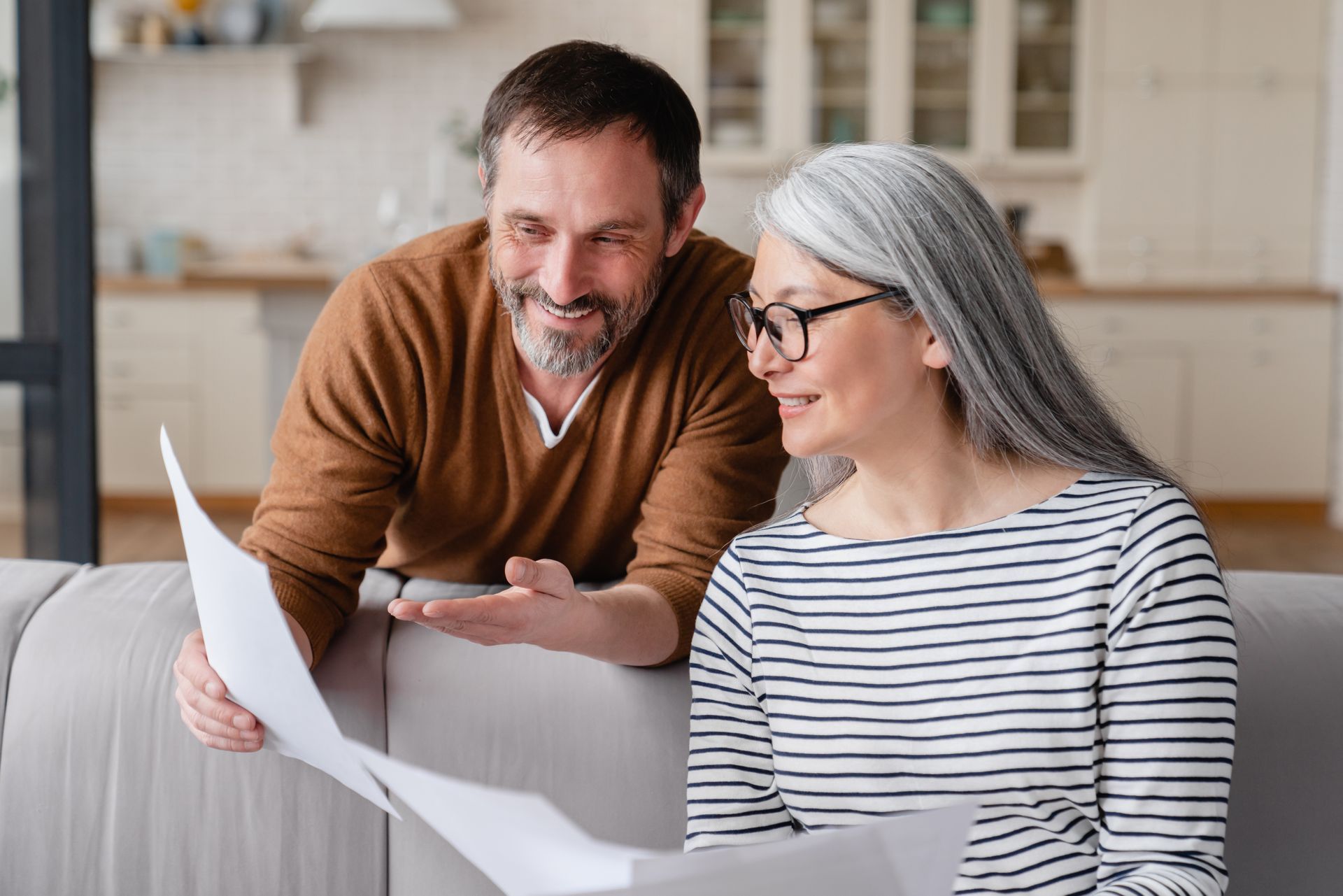 Couple reviewing documents on a couch, man points, woman wears glasses, light-filled room.
