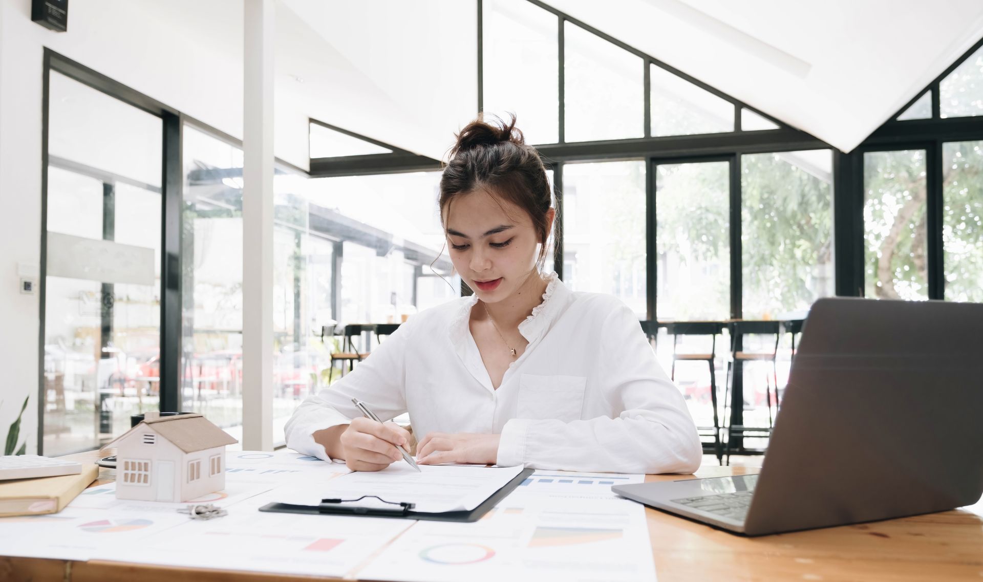 Woman in white shirt writing at desk with laptop, model house, and papers.