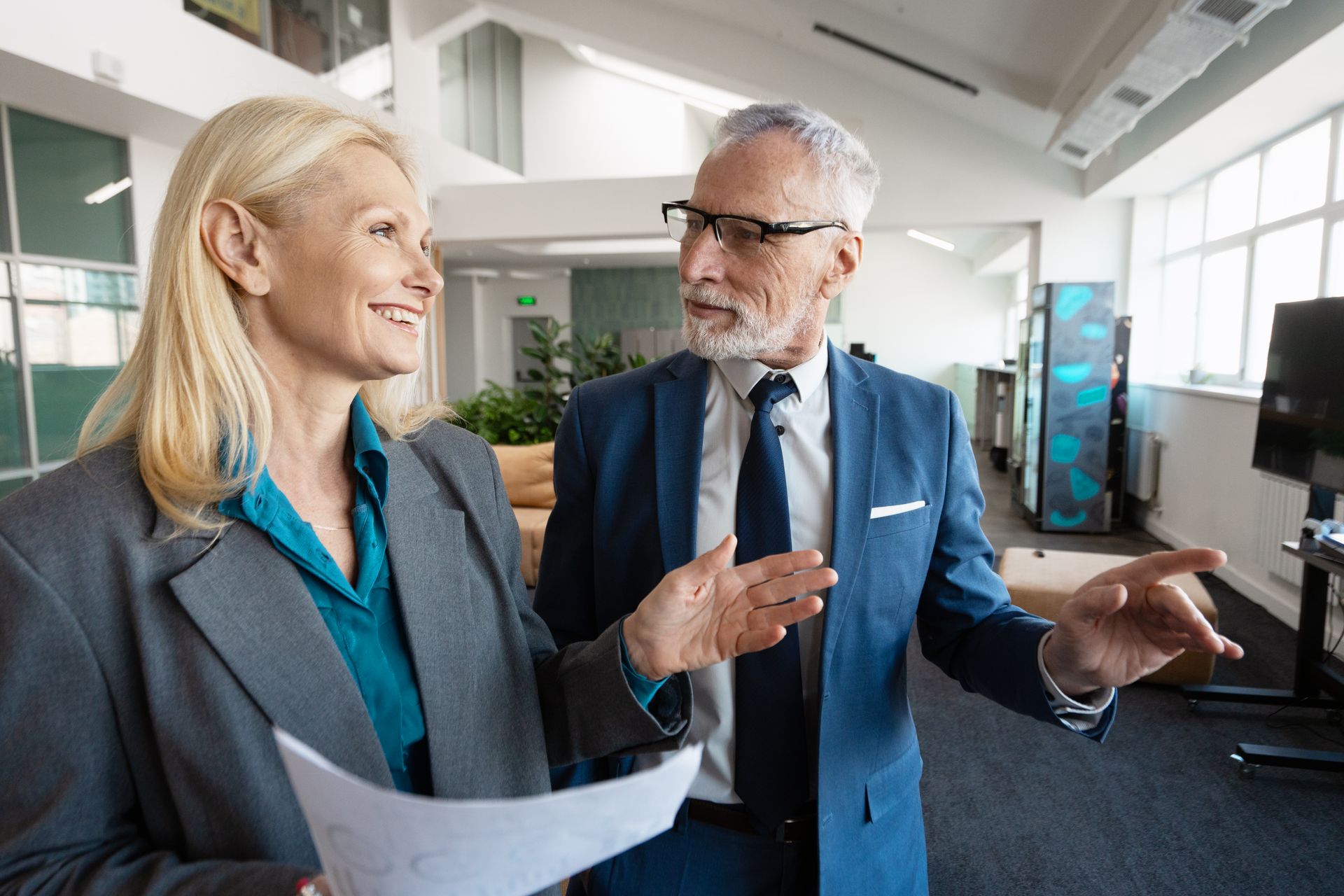 Two people in business attire, looking at each other, one holding papers, inside a modern office.