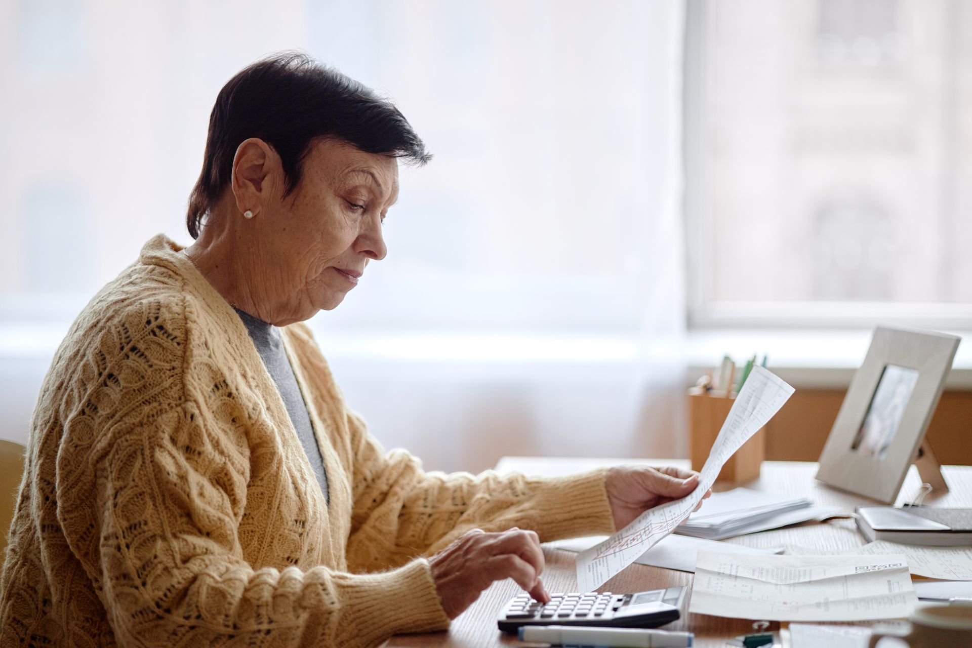 Woman in yellow sweater using calculator, reviewing papers at a desk near a window.