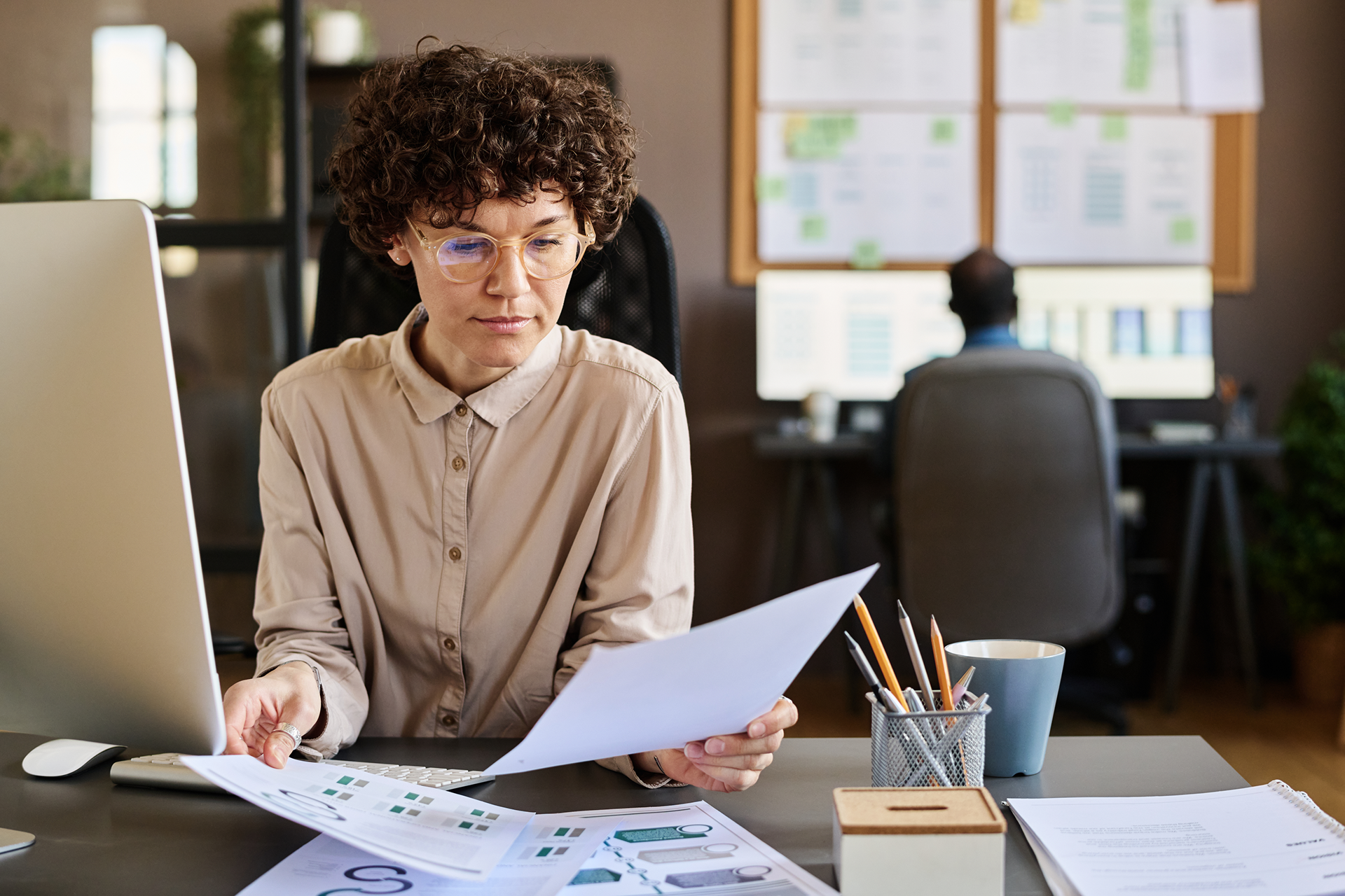 Woman with glasses reviews paperwork at a desk, holding a tablet and using a calculator, with a bicycle in the background.