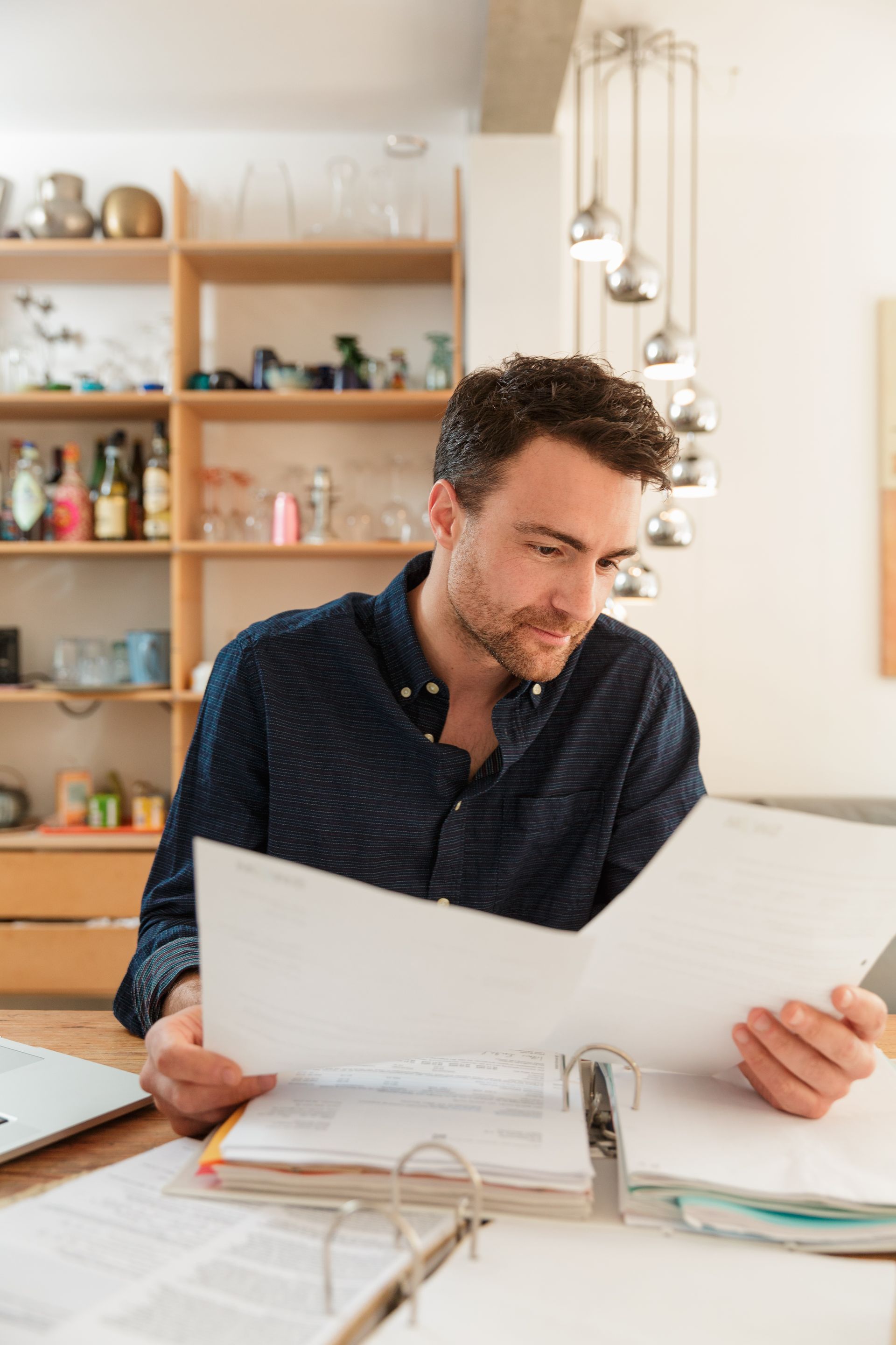 Man reviews documents at a desk, looking thoughtful. Bright room, wooden shelves in the background.