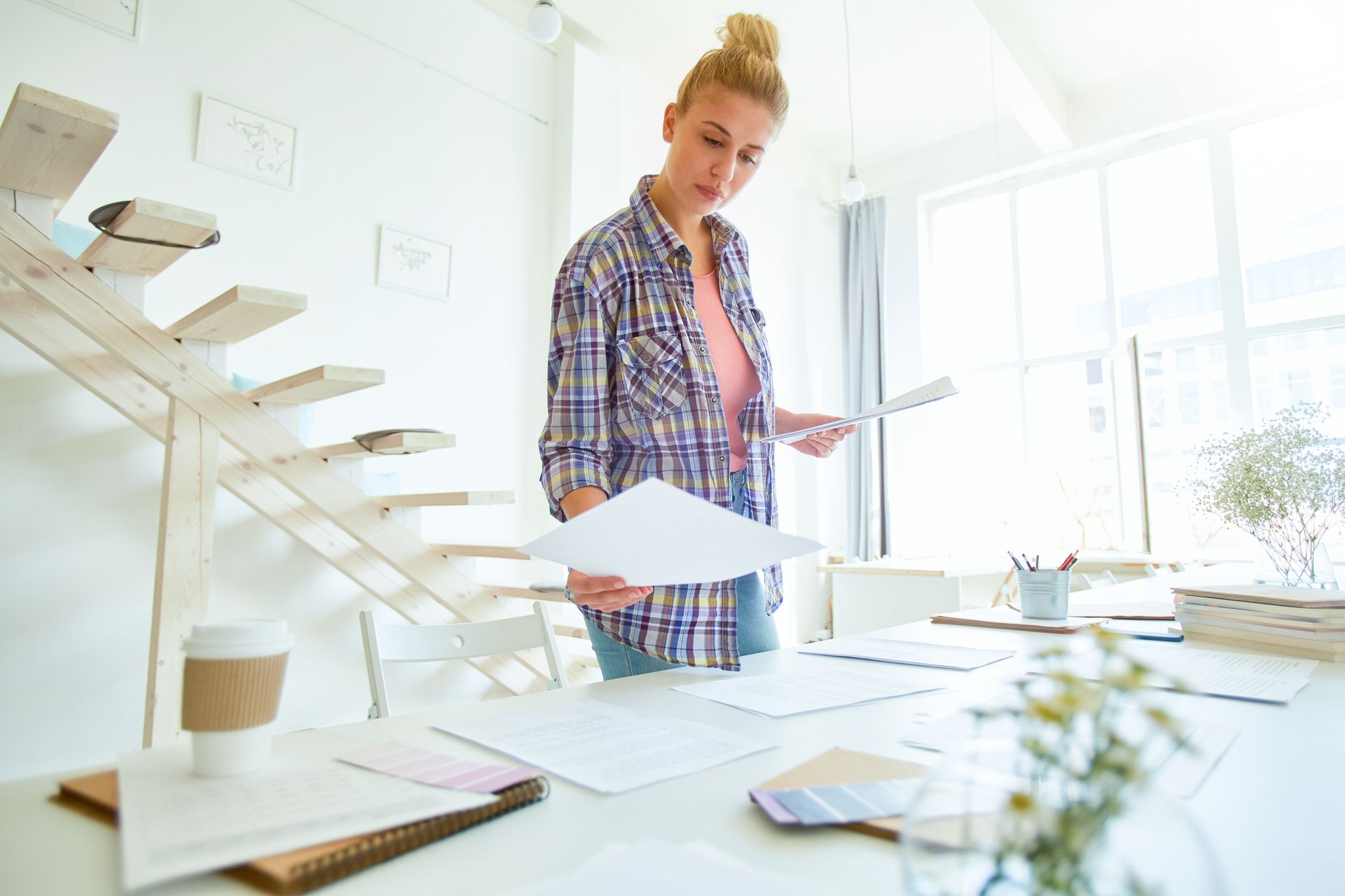 Woman standing at a table, reviewing papers, bright room, bun hairstyle, plaid shirt.
