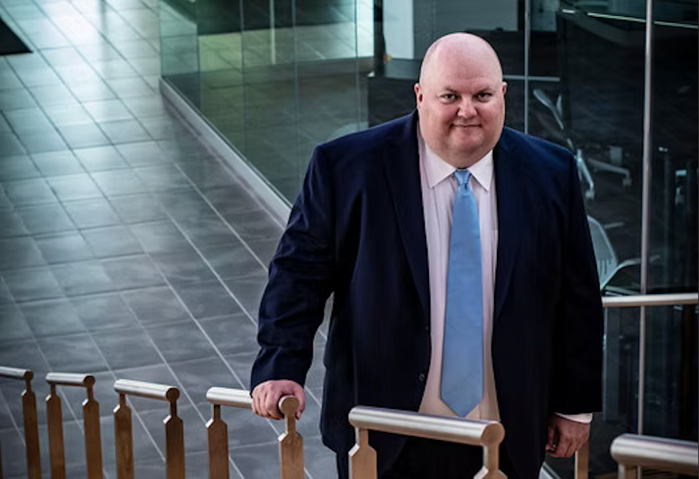 Man in a blue suit stands on a staircase, smiling.