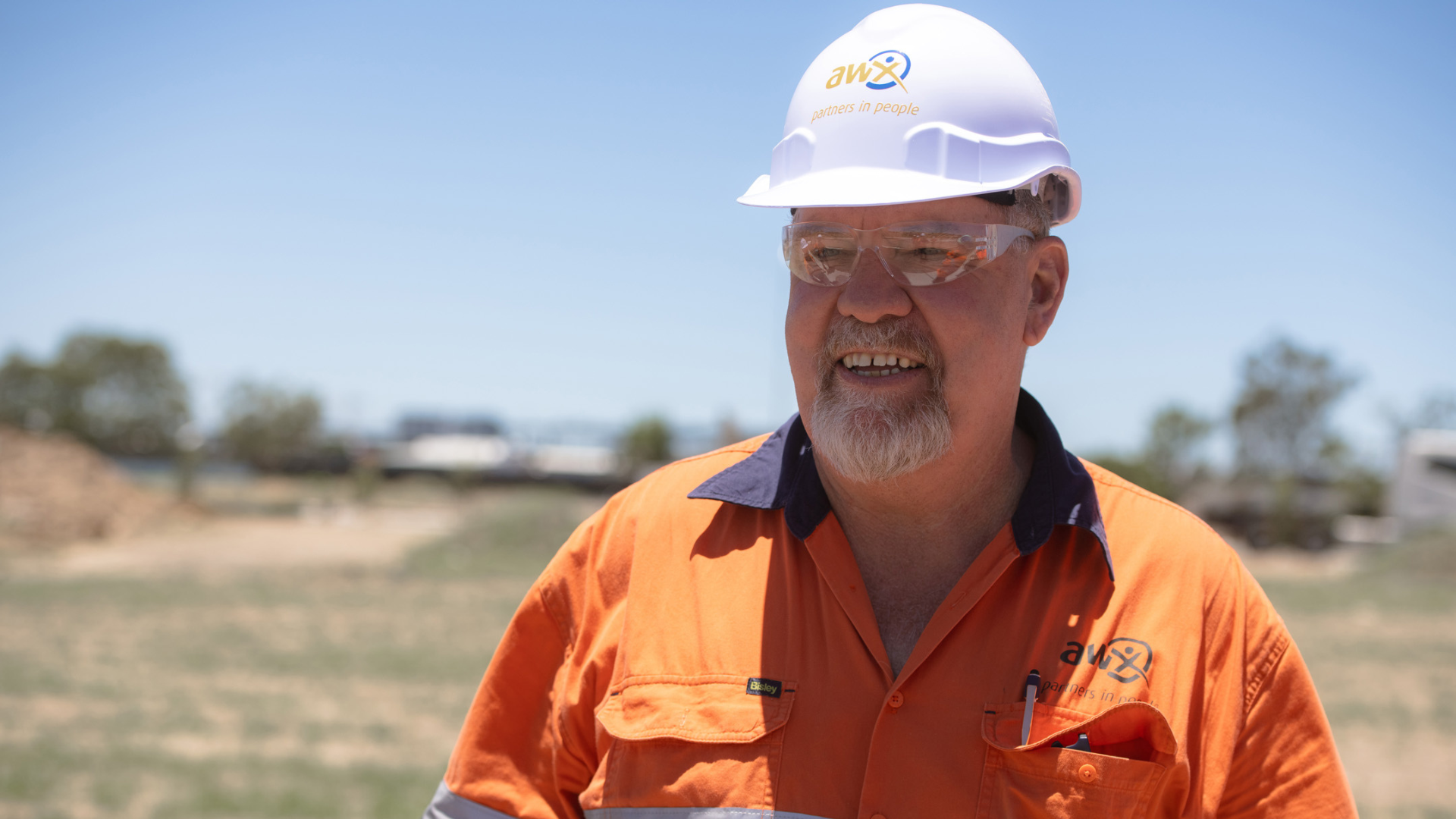 Man in hard hat and orange work shirt smiles outdoors.