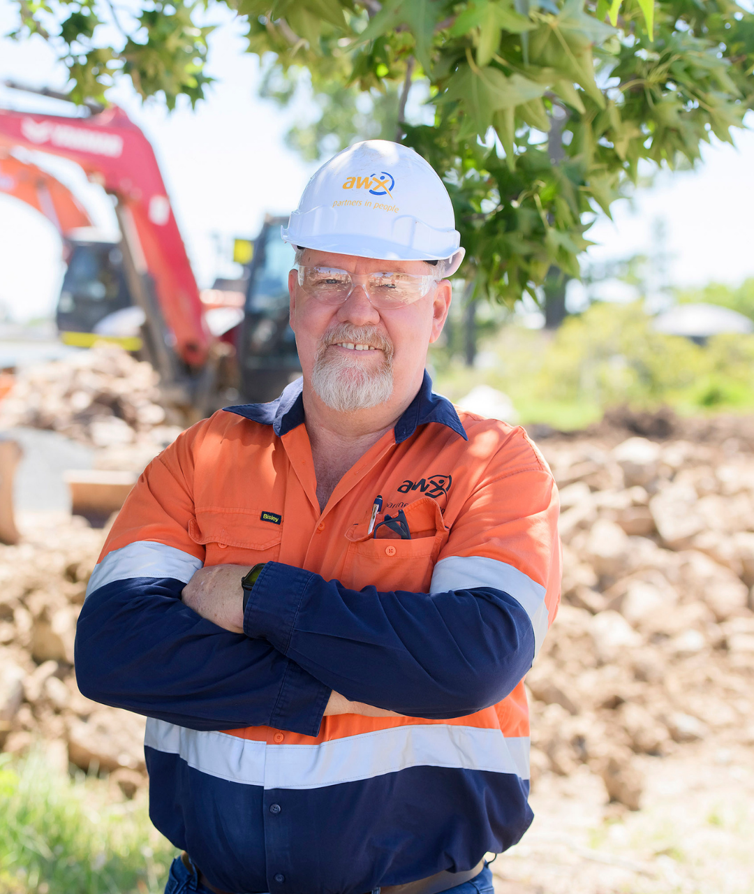 Man in workwear and hardhat, arms crossed, smiling at camera; construction site background.