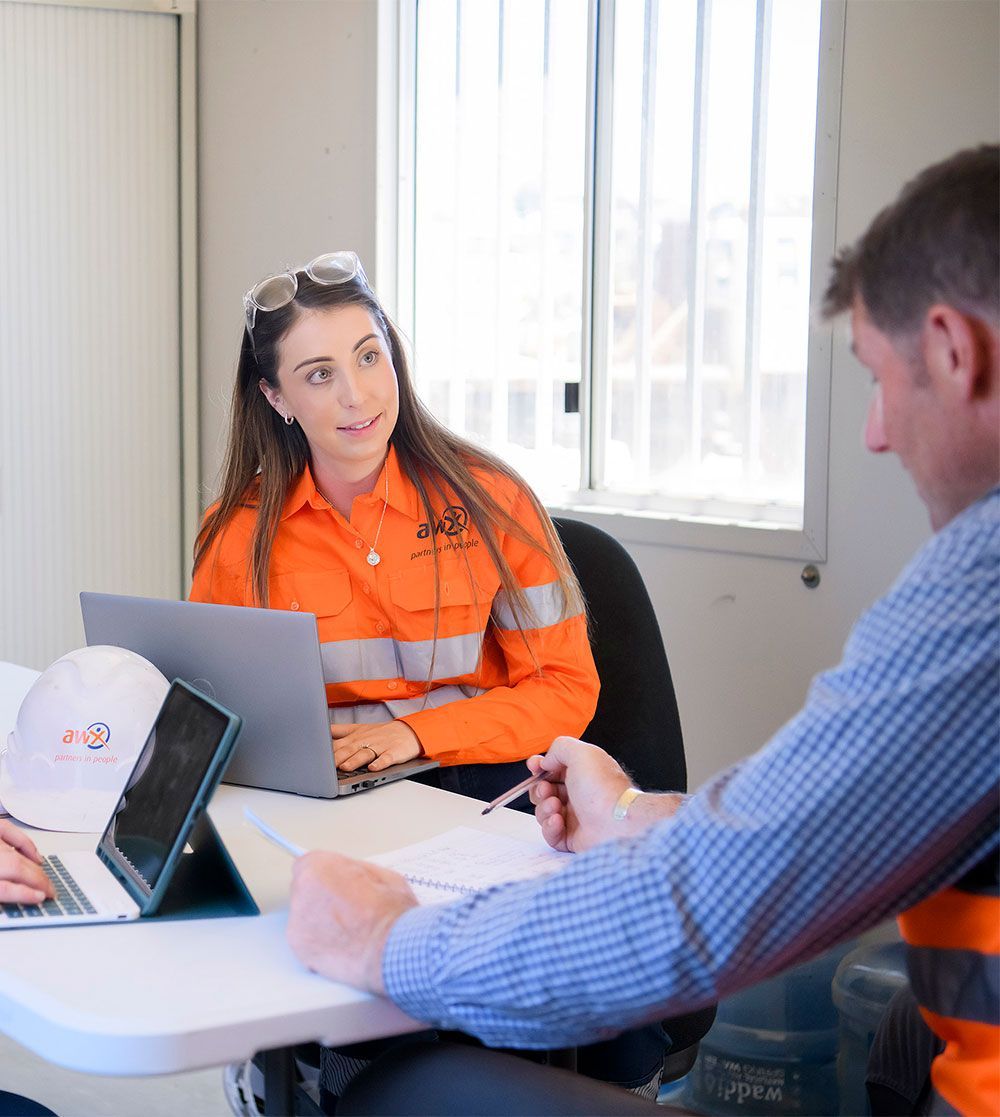 Woman in orange workwear, laptop, talking to a man in a room with a window, possibly a work meeting.