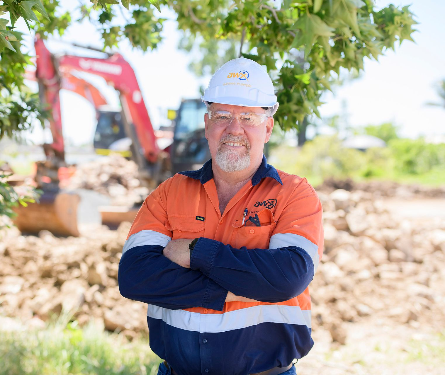 Man in construction workwear, arms crossed, smiling. Hard hat, excavator in background.