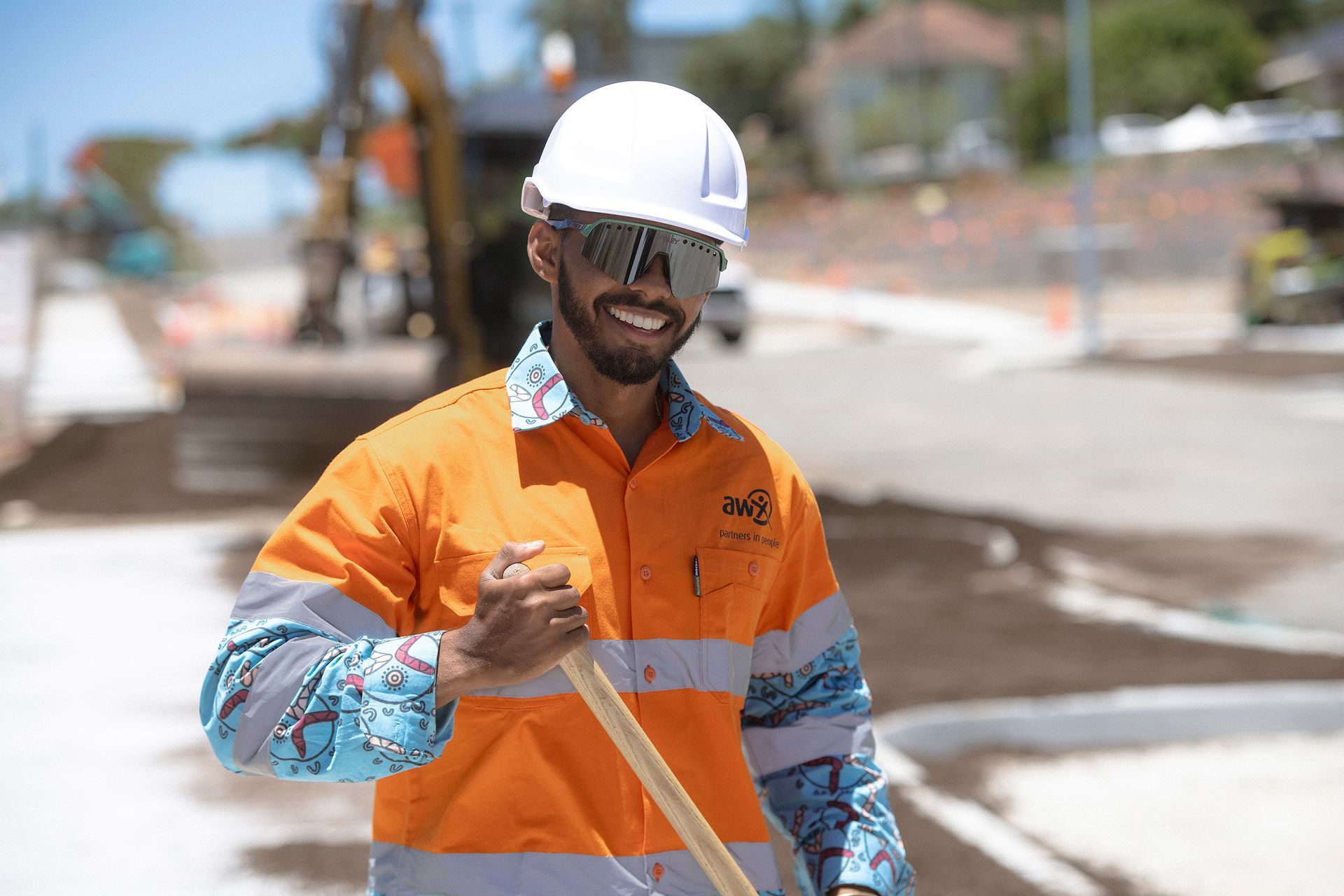 Construction worker smiling, holding a tool, wearing a white helmet and reflective orange shirt.