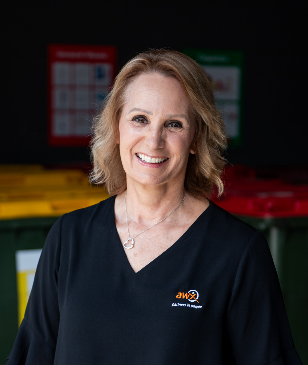 Woman in black top smiles, standing in front of recycling bins.