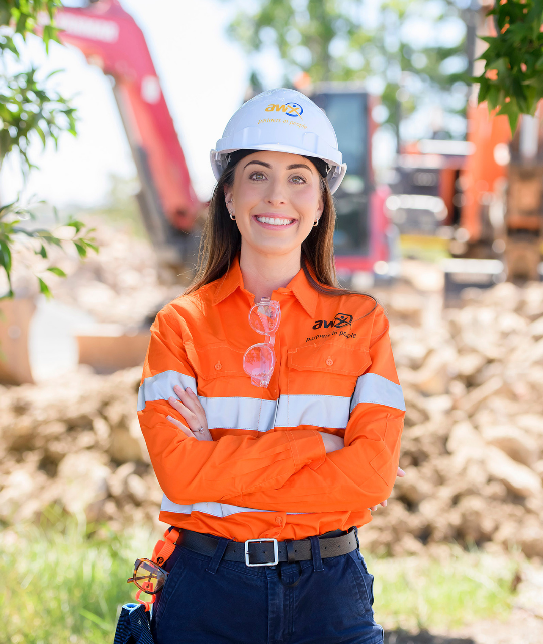 Woman in safety gear, smiling with arms crossed, in a construction site with machinery.