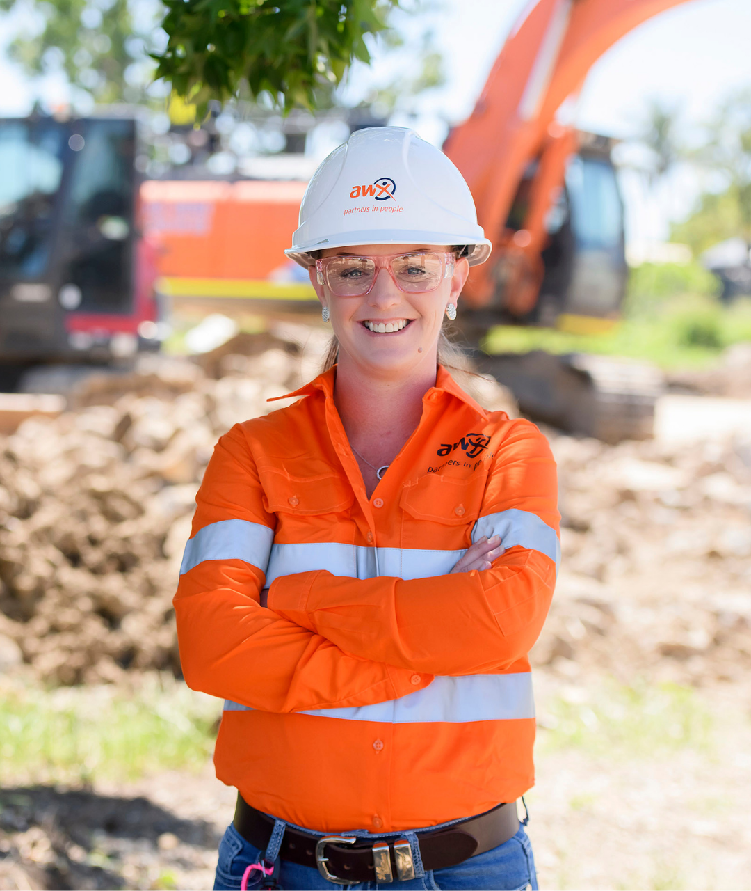 Woman in construction attire smiles, arms crossed. Orange safety vest, white hard hat. Construction site background.
