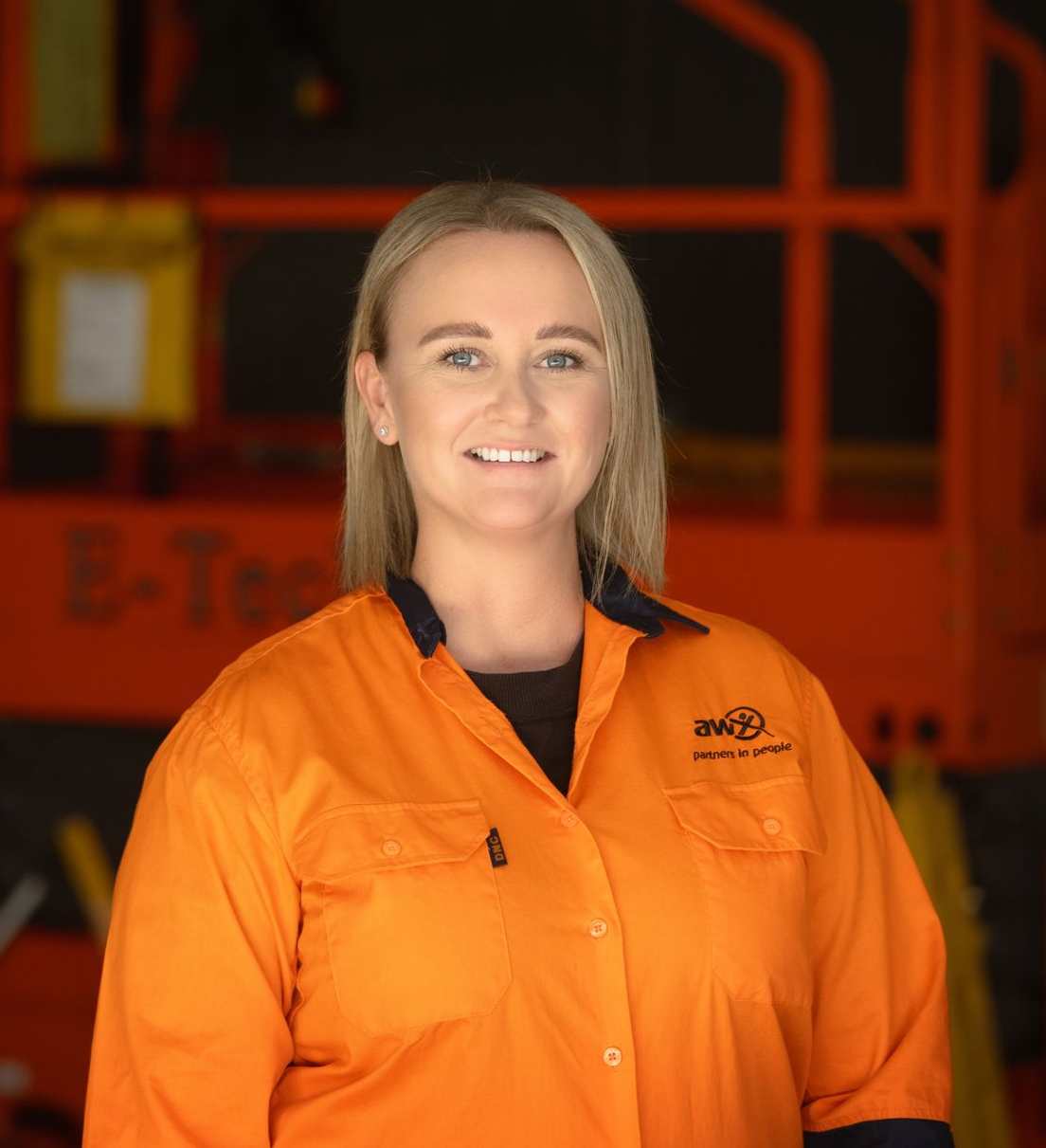 Man in orange and blue work shirt smiles, arms crossed, leaning against wooden post. Outdoors setting.