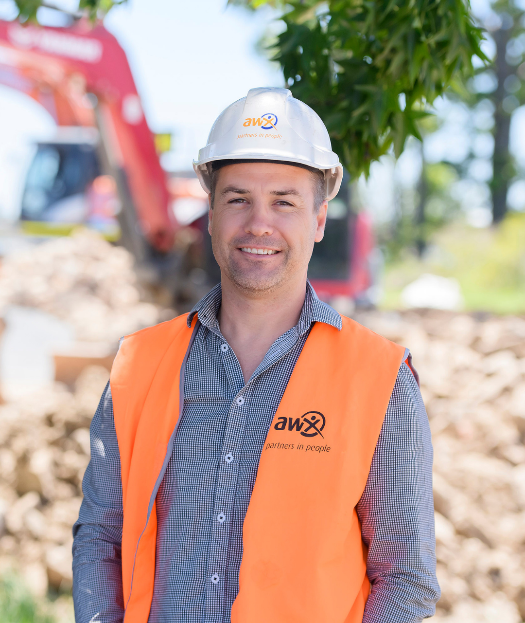 Man in hard hat and orange vest smiles, construction site with excavator in background.