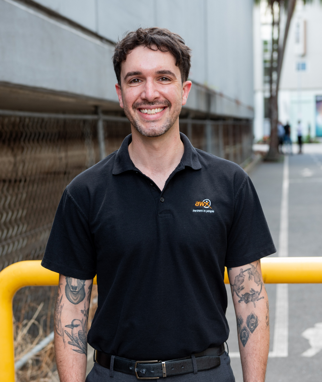 Man smiling, wearing a black polo shirt with a logo, standing near a yellow barrier and a building.