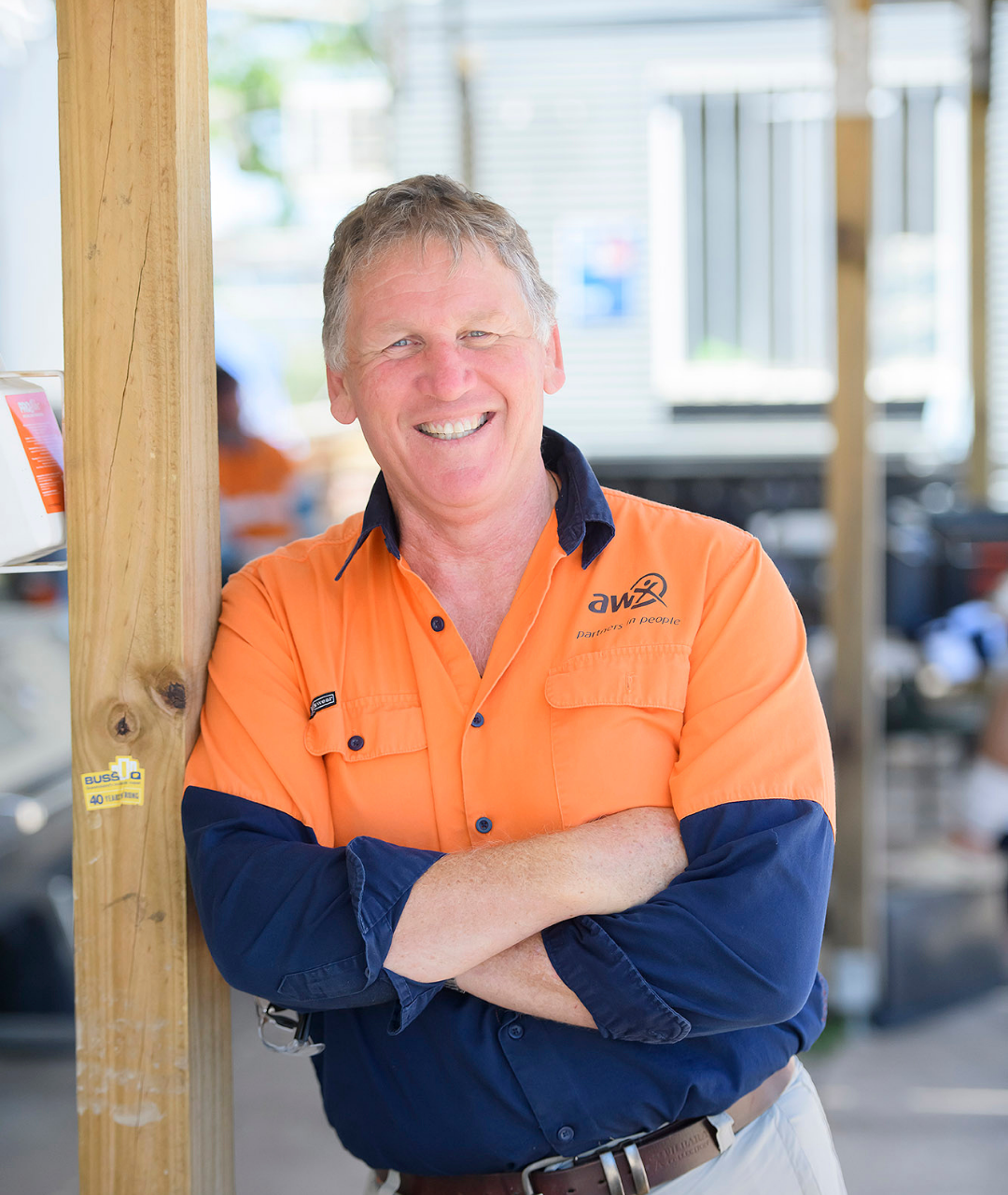 Man in orange and blue work shirt smiles, arms crossed, leaning against wooden post. Outdoors setting.