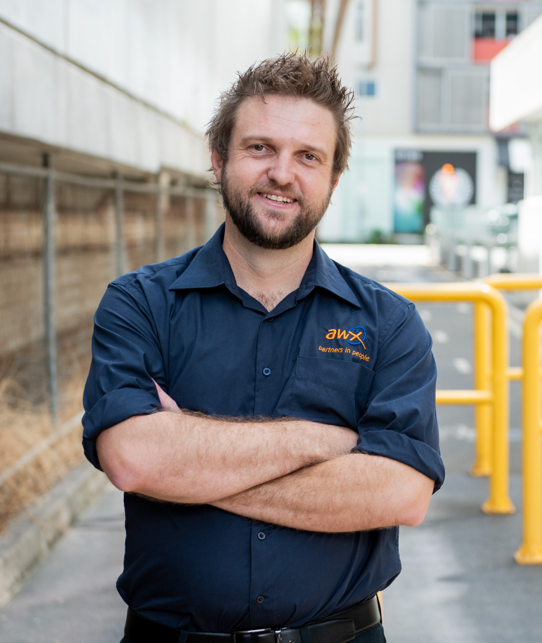 Man with beard, blue shirt, arms crossed, smiling, outdoors.