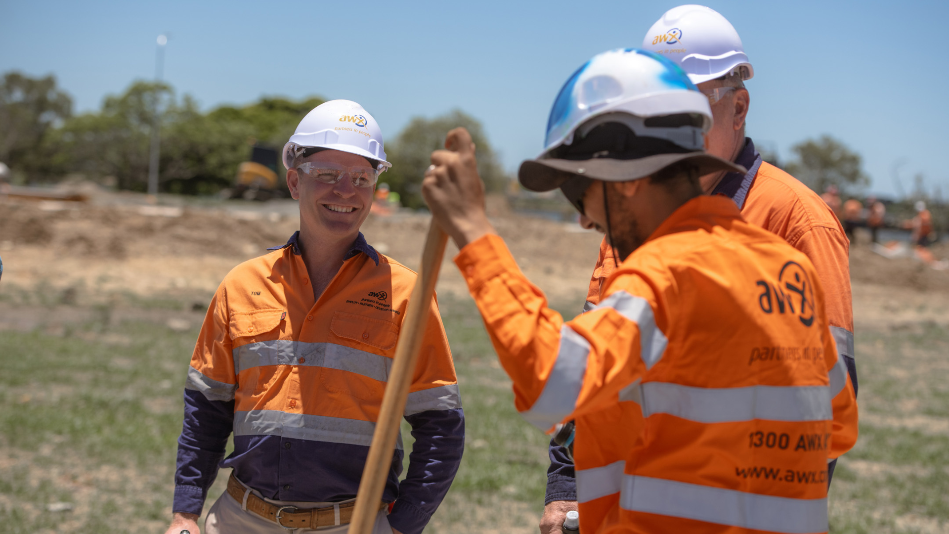 Three construction workers in orange safety gear on a sunny worksite. One holds a shovel, smiling.