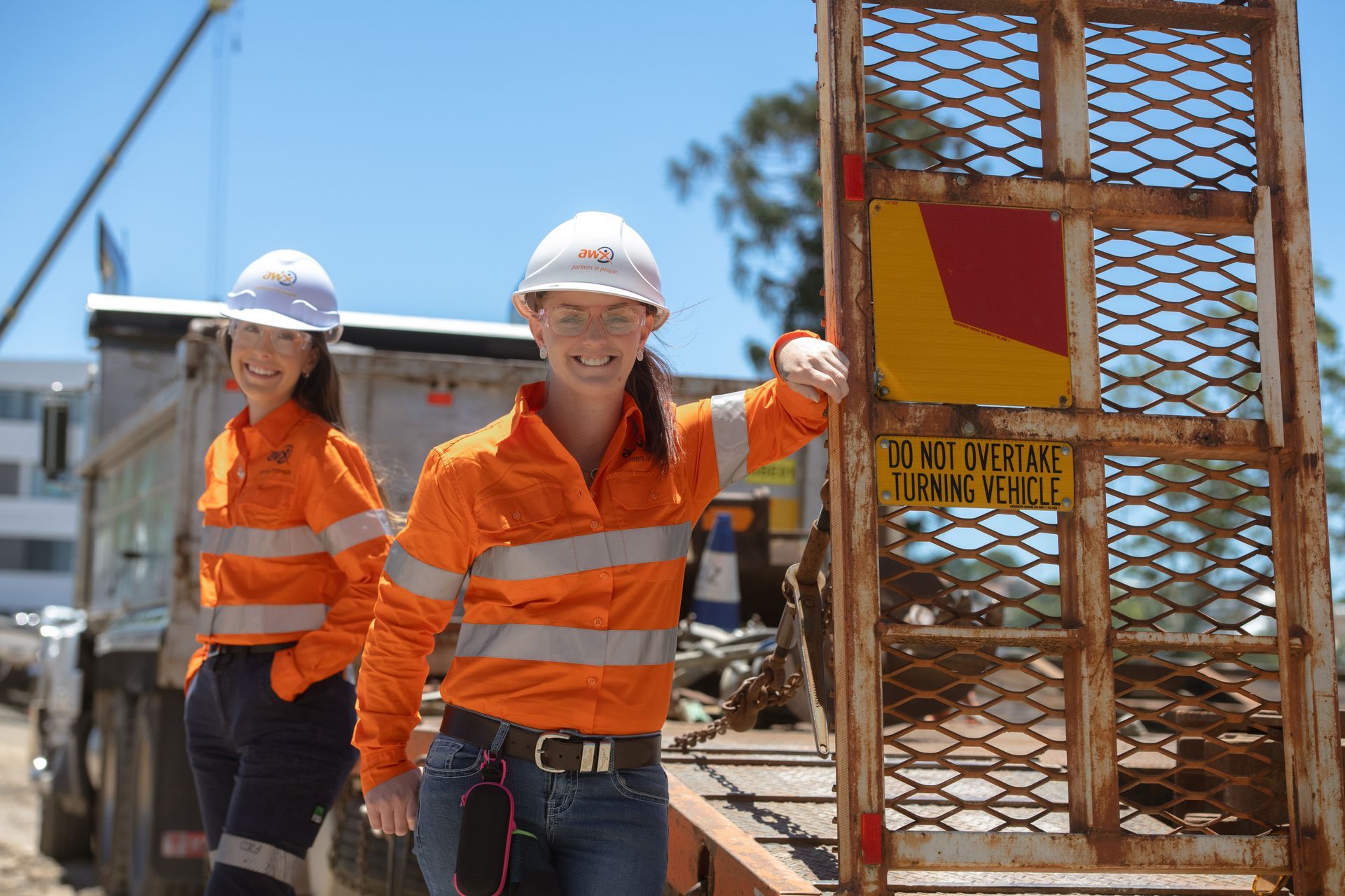 Two women in orange safety vests and hard hats smile at camera, next to a truck.